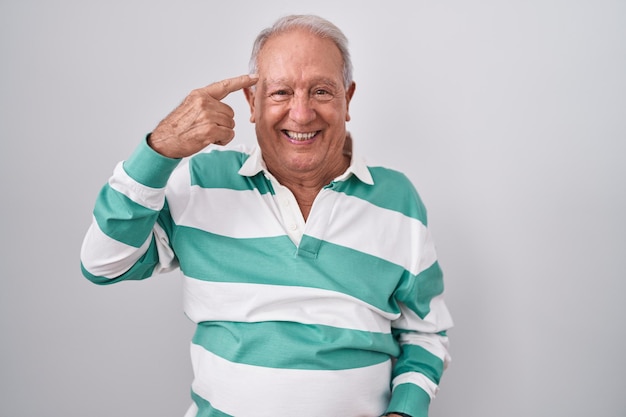 Senior man with grey hair standing over white background smiling pointing to head with one finger, great idea or thought, good memory