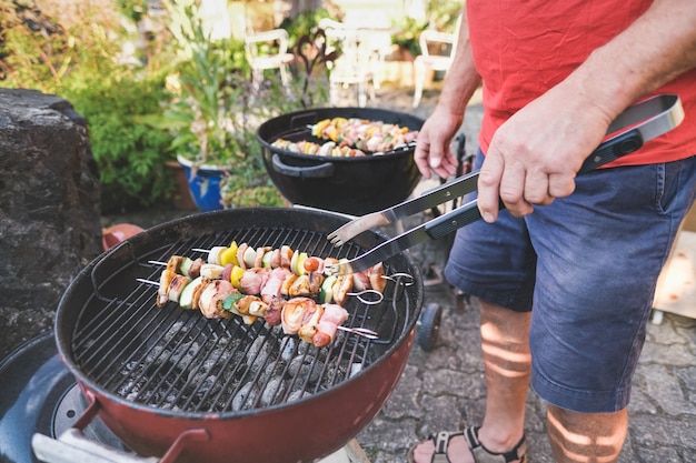 Photo senior man with grill tongs turns the meat and vegetable skewers on the grill - barbecue party in the garden