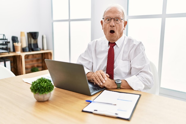 Senior man working at the office using computer laptop afraid and shocked with surprise and amazed expression fear and excited face