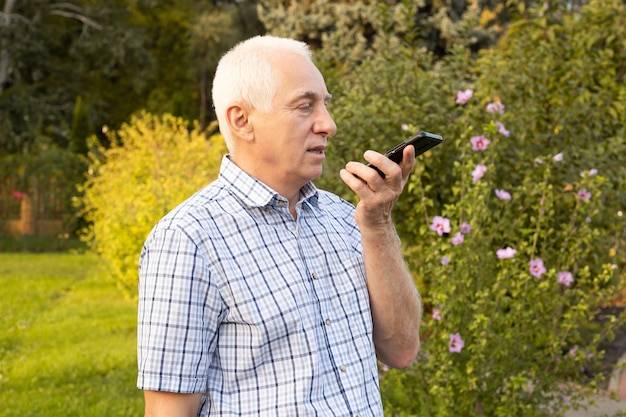 Senior mature old man using mobile phone with voice search in green park, modern technologies