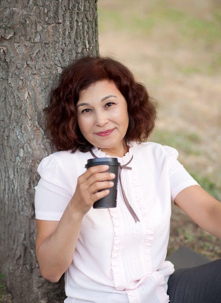 Senior middle age woman sitting in a park outdoor and drinking coffee from paper cup