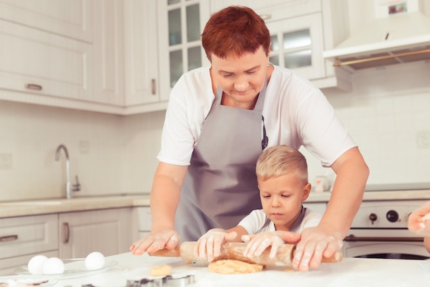 Senior nanny or grandmother helping child to roll out dough