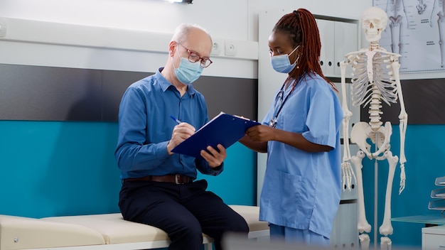 Senior patient signing medical form at checkup with nurse during coronavirus pandemic. Old man doing consultation appointment with specialist, receiving prescription treatment.