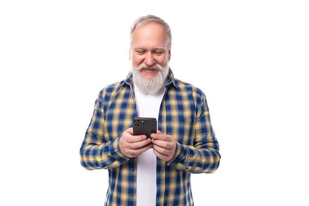 Senior pensioner grayhaired man with a beard in a shirt masters a smartphone on a white background