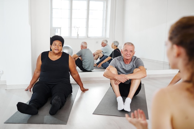 Senior people doing relaxation exercises on the floor and listening to the coach during gymnastics i...