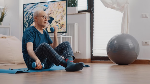 Senior person using dumbbells to exercise and train arms muscles, sitting on yoga mat. Retired man lifting weights and training with fitness equipment. Pensioner stretching and exercising