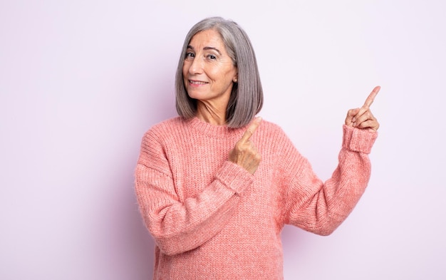 Senior pretty woman smiling happily and pointing to side and upwards with both hands showing object in copy space