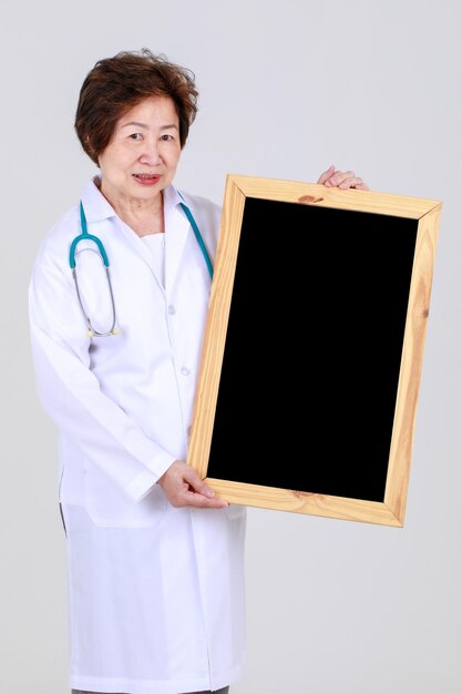 Senior professional female physician on medical gown with stethoscope holding square wooden blackboard with smile to express health care info and disease treatment from elderly doctor at hospital.