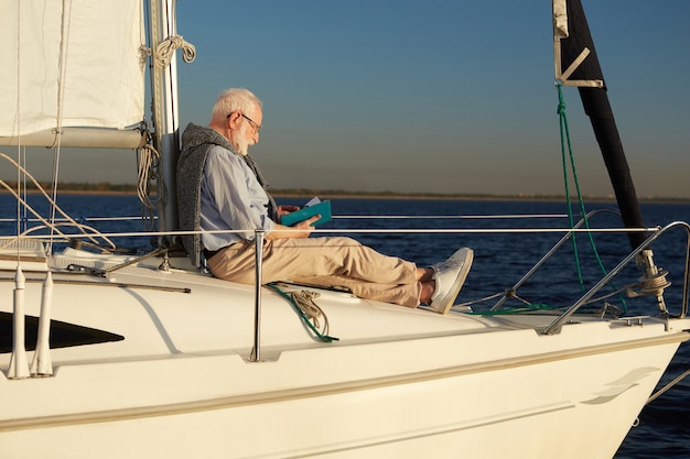 Senior relaxed man reading book while sitting on the side of sailboat or yacht deck floating in the