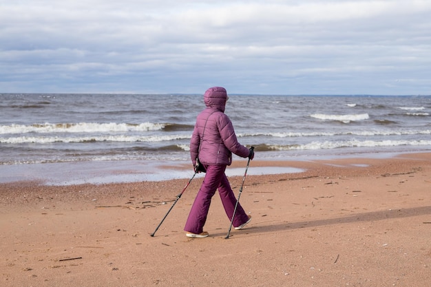 Senior retired woman hiking on the seaside on the sand Active fitness and healthy lifestyle Female training on the beach Nordic walking with poles Autumn season at the seaside Sporty couple