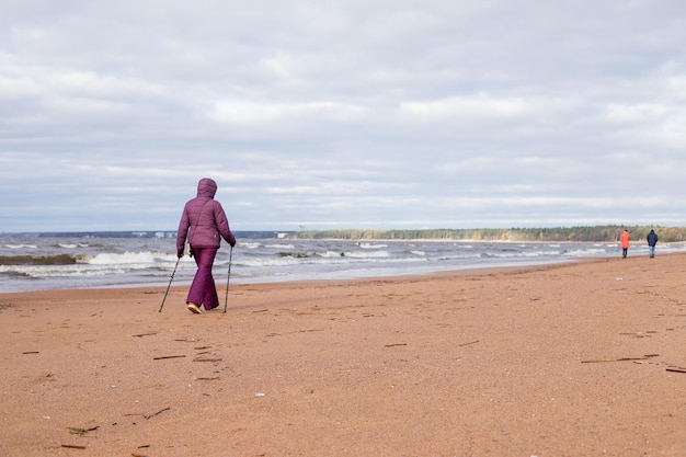 Senior retired woman hiking on the seaside on the sand Active fitness and healthy lifestyle Female training on the beach Nordic walking with poles Autumn season at the seaside Sporty couple