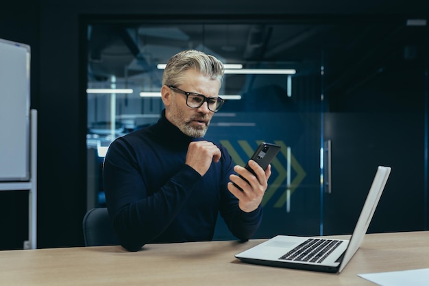 Senior serious grayhaired male businessman sitting in the office at the desk and using a mobile