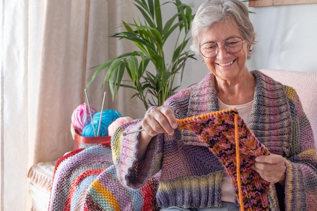 Senior smiling woman sitting on sofa at home while knitting and looking at her work in progress