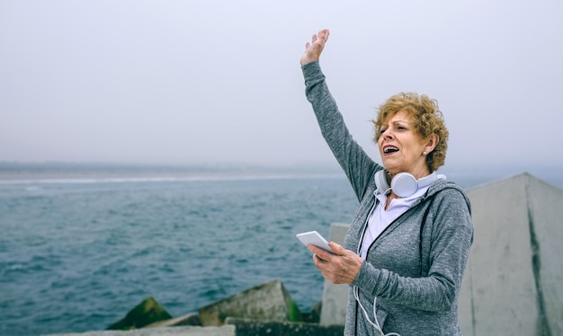 Senior sportswoman using smartphone and waving by sea pier