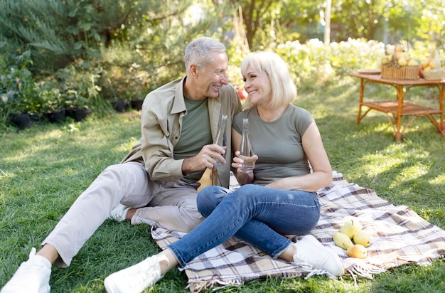 Senior spouses clinking bottles with fresh water having picnic and sitting on blanket in their