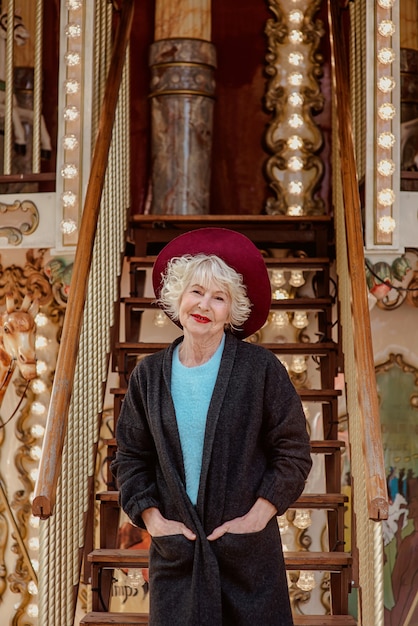 Photo senior stylish woman in dark grey coat hat and with grey hair standing by the carousel smiling