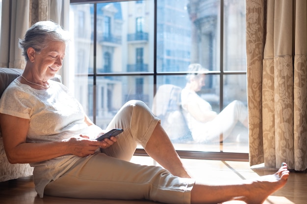 Senior traveler woman in hotel room sitting on the floor consulting her smart phone to organize a city tour