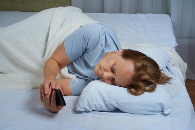 Senior woman in blue pajamas using mobile phone while lying in bed suffering from insomnia.