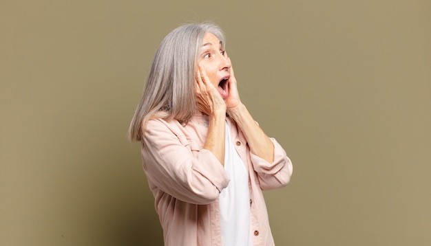 Senior woman feeling happy, excited and surprised, looking to the side with both hands on face