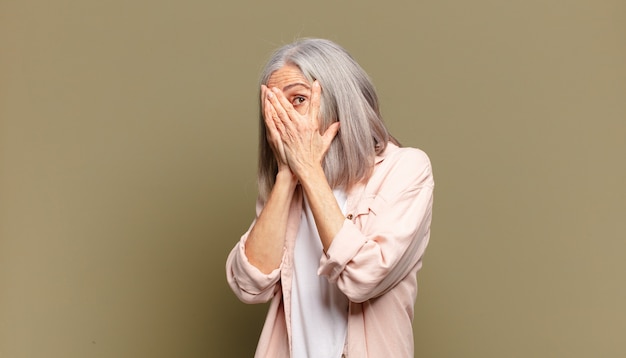 Senior woman feeling scared or embarrassed, peeking or spying with eyes half-covered with hands