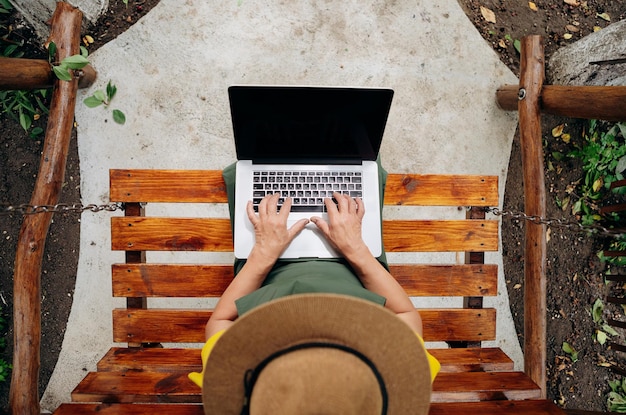 Senior woman gardener in a hat is watching something on a laptop computer and making a video call in the yard while outdoors