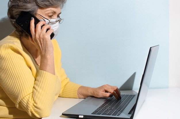 Senior woman in glasses and medical mask calling by phone. Elderly woman using laptop at home. Concept for online consultation, remote working, new normal.