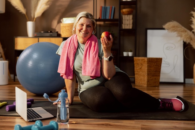 Senior woman granny sitting on yoga matholding apple in arm blue dambbells and bottle with water