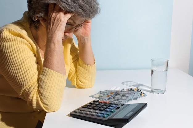 Senior woman holding her head. Pensioner sitting at the table with medicines and money. Concept of depression, drug price, treatment cost