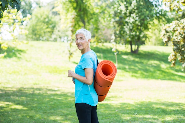 Senior woman holds fitness mat on her back in the park and preparing for exercise