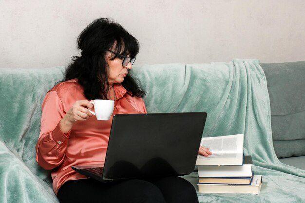 senior woman at home on the sofa reading book holding laptop on her lap and cup of coffee in her hand selfeducation at any age