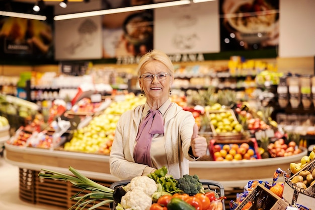 A senior woman is purchasing at the supermarket and giving thumbs up while smiling at the camera