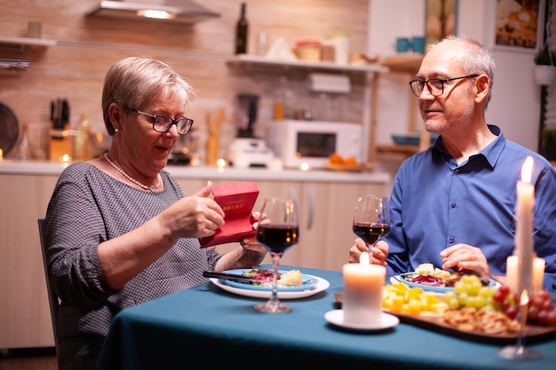 Senior woman looking surprised at gift box from husband during dinner in kitchen. Happy cheerful elderly couple dining together at home, enjoying the meal, celebrating their marriage , surprise holida