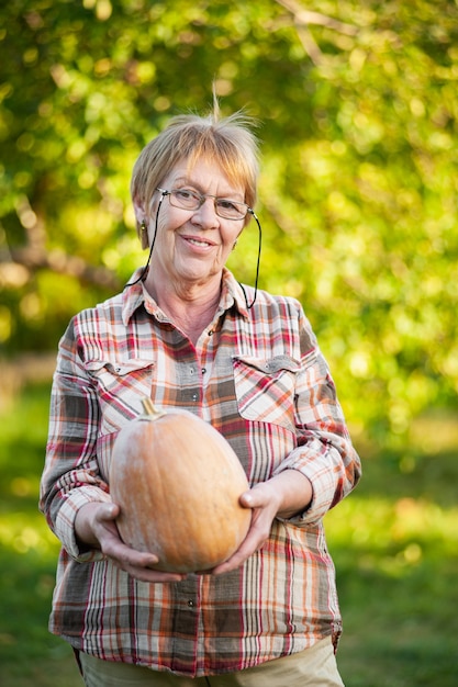 Senior woman in a plaid shirt holds a pumpkin in the garden.