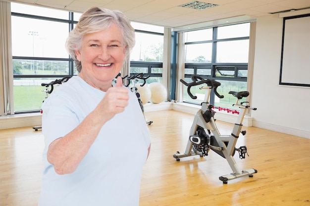 Senior woman showing thumbs up against spinning exercise bikes in gym room