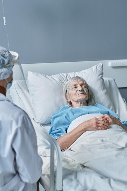 Senior woman sleeping in the hospital bed with doctor observing her at the ward