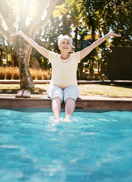 Senior woman smile and feet in swimming pool in relax for summer vacation swim or holiday joy in the outdoors Happy elderly female with arms stretched out in happiness for relaxation by water pool