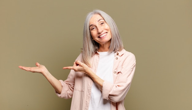 Senior woman smiling cheerfully and pointing to copy space on palm on the side, showing or advertising an object