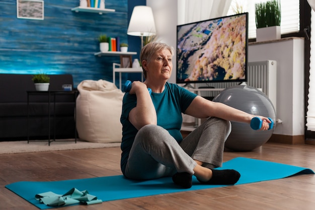 Senior woman in sportwear warming up arm muscles practicing wellness workout using dumbbells pension...