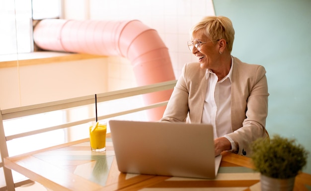 Senior woman using mobile phone while working on laptop and drinking fresh orange juice in the cafe