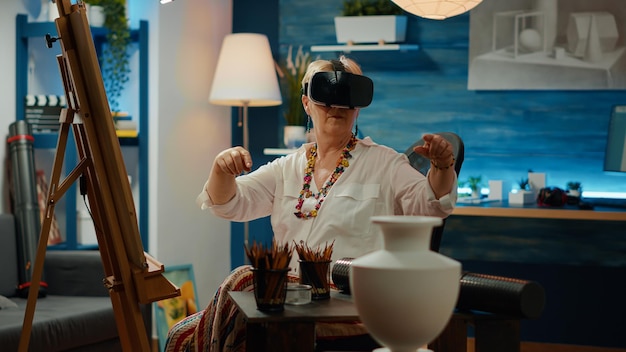 Senior woman using vr glasses with augmented reality to create drawing of vase for art and craft skills. Interactive 3d simulation on wireless headset to draw artwork sketch and masterpiece.