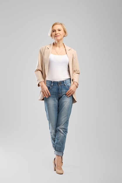 Senior woman in white tank top beige jacket and boyfriend jeans walking in studio over grey background