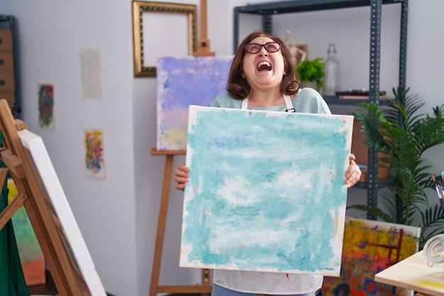 Senior woman with glasses holding canvas at art studio angry and mad screaming frustrated and furious, shouting with anger looking up.