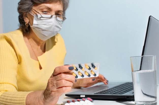 Senior woman with glasses and medical mask reading instructions of medicine, selective focus. Elderly woman using laptop at home. Concept for online consultation with doctor, new normal.