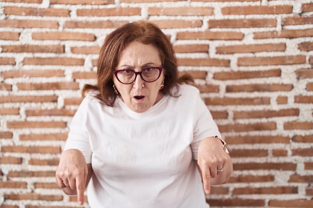 Senior woman with glasses standing over bricks wall pointing down with fingers showing advertisement, surprised face and open mouth