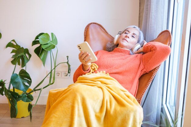 Senior woman with gray hair sitting relaxed listening to music from her smartphone and headphones Concept retirement rest music