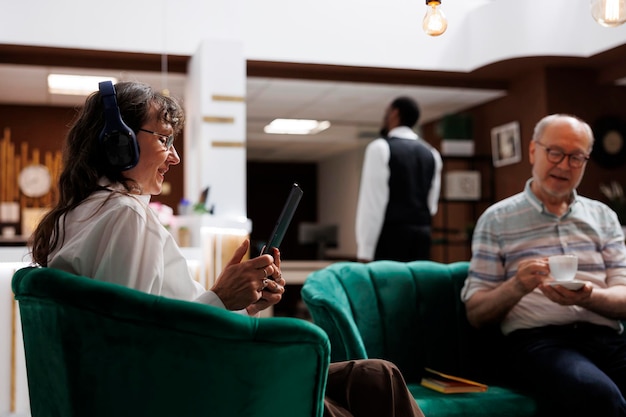 Senior woman with headphones sitting on sofa in hotel lobby confirming reservation on device Male customer holding a cup while elderly lady with wireless headset uses her tablet in lounge area