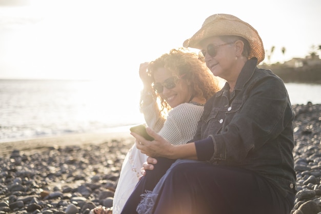 Senior woman with her young daughter relaxing on beach. Mother showing mobile phone to daughter and smiling. Old woman sharing media content with her elder daughter on beach on a bright sunny day