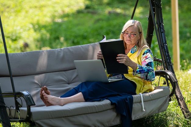 Photo senior woman with laptop and documents working in garden on rocking couch green home office concept