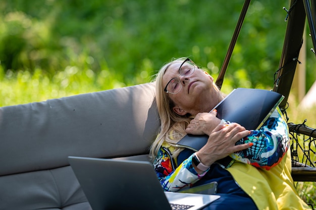 Senior woman with laptop tired relaxing in garden on rocking sofa leisure and vacation concept