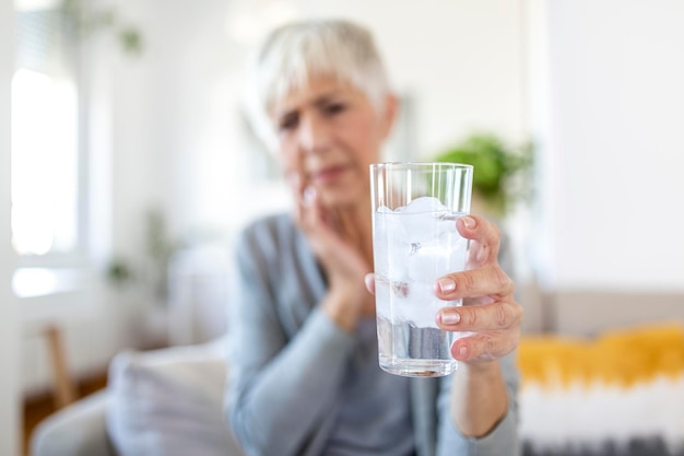 Senior woman with sensitive teeth and hand holding glass of cold water with ice. Healthcare concept. Mature woman drinking cold drink, glass full of ice cubes and feels toothache, pain